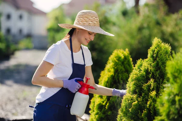 Mulher jardineiro bonita em chapéu de palha asperge plantas de um pulverizador de jardim — Fotografia de Stock
