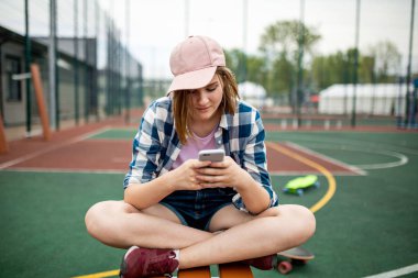 Kareli gömlek giymiş bir sarışın kız pembe kap ve kot şort bağdaş kurup spor alanında oturup ve telefon arıyor. Yeşil bir longboard arka planda olduğunu.