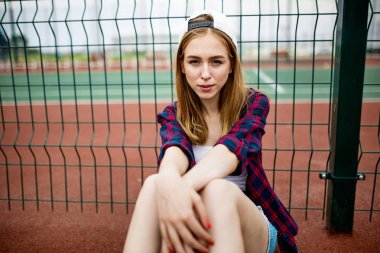 Çok gülümseyen bir sarışın kız kareli gömlek, beyaz kap ve kot şort giyen, spor sahasında leaned guardlattice üzerinde oturuyor. Spor ve serin tarzı.