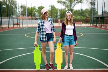 Damalı gömlek, şapka ve kot şort giyen iki güzel gülümseyen sarışın kız ellerinde parlak longboards ile spor alanında duruyor. İyi uyum içinde güzel bacaklar. Spor ve serin tarzı