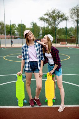 Damalı gömlek, şapka ve kot şort giyen iki güzel gülümseyen sarışın kız ellerinde parlak longboards ile spor alanında duruyor. İyi uyum içinde güzel bacaklar. Spor ve serin tarzı