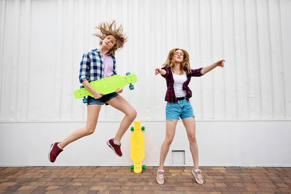 Duas lindas garotas loiras vestindo camisetas quadriculadas e shorts jeans estão pulando e dançando com longboards brilhantes. Meninas jovens estão cativando e se divertindo . — Fotografia de Stock