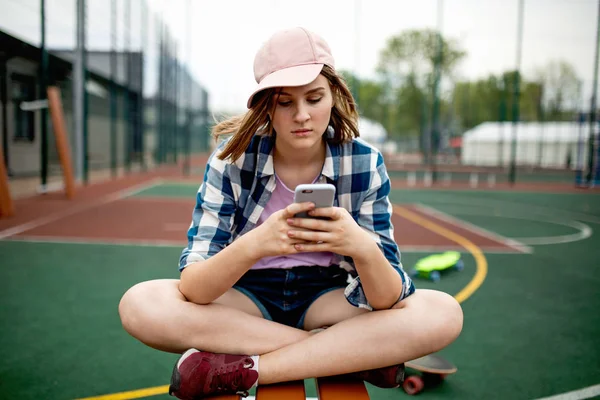 En ganska blond flicka klädd i rutig skjorta, rosa mössa och denim shorts sitter skräddarställning på idrottsplanen och tittar på telefonen. En grön longboard är i bakgrunden. — Stockfoto
