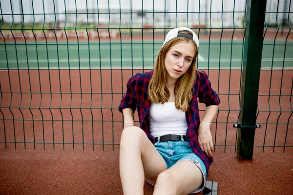 Ein hübsches blondes Mädchen mit kariertem Hemd, weißer Mütze und Jeanshose sitzt angelehnt an das Gitter auf dem Sportplatz. Sport und cooler Stil. — Stockfoto