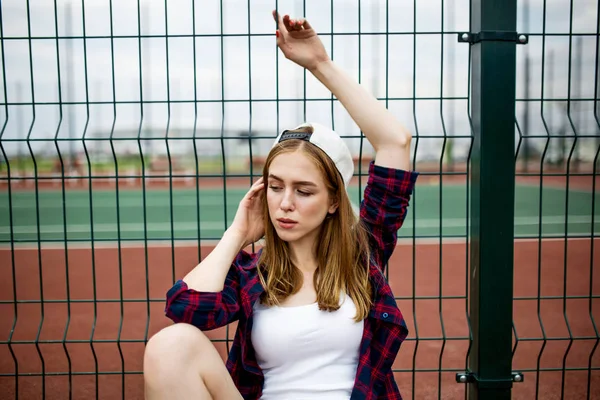 一个穿着格子衬衫、白帽和粗斜纹棉布短裤的金发女孩正坐在运动场上的护栏上, 双手举过头顶。运动和清凉风格. — 图库照片