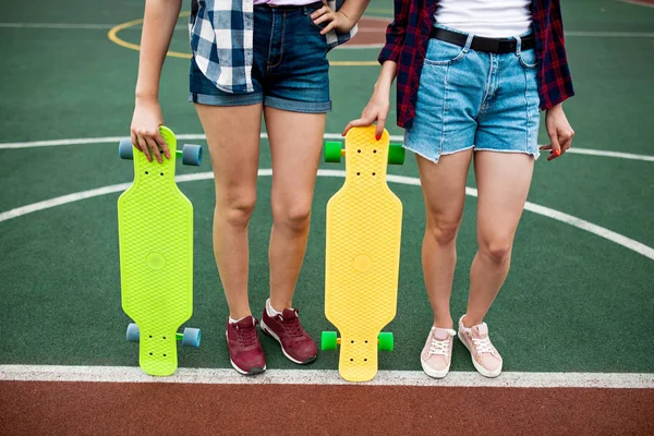 체크 무늬 셔츠와 데님 반바지를 입은 두 소녀가 밝은 롱보드를 손에 들고 운동장에 서 있다. 아름 다운 다리에 딱 맞는. — 스톡 사진