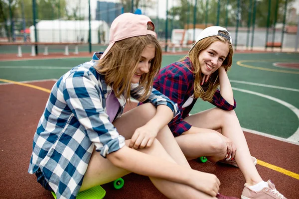 Két csinos szőke lány visel kockás ingek, sapkák és farmer rövidnadrág ül a fényes longboards a sportsfield. Szép lábak, jó illeszkedést. Sport-és hidegstílus. — Stock Fotó