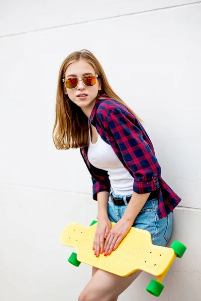 一个戴着太阳镜、格子衬衫和牛仔短裤的金发女孩站在灰色的墙壁前，在她面前拿着一块黄色的长板。酷炫风格. — 图库照片