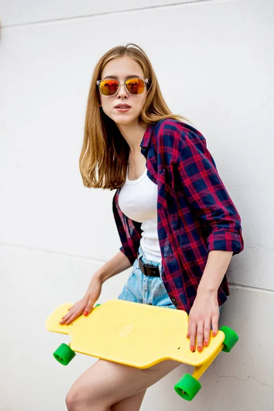 Uma linda garota loira usando óculos de sol, camisa quadriculada e shorts de ganga está em frente à parede cinza e segurando um longboard amarelo na frente dela. Estilo legal . — Fotografia de Stock