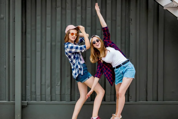 两个漂亮的微笑的金发女孩穿着格子衬衫，帽子和牛仔短裤在长板上的黑色墙前平衡，玩得很开心。运动和酷炫的风格. — 图库照片