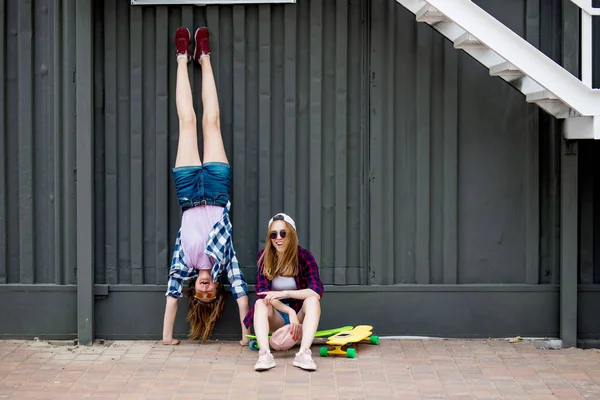 Duas garotas loiras sorridentes vestindo camisetas quadriculadas, bonés e shorts de ganga estão equilibrando na frente da parede preta nos longboards e se divertindo de cabeça para baixo. Esporte e legal — Fotografia de Stock