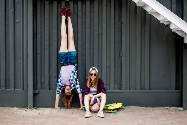 Duas garotas loiras sorridentes vestindo camisetas quadriculadas, bonés e shorts de ganga estão equilibrando na frente da parede preta nos longboards e se divertindo de cabeça para baixo. Esporte e legal — Fotografia de Stock