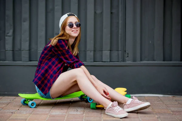 Uma linda garota loira usando óculos de sol, camisa quadriculada e shorts de ganga está sentada nos painéis de madeira brilhantes em frente à parede preta. Estilo legal — Fotografia de Stock