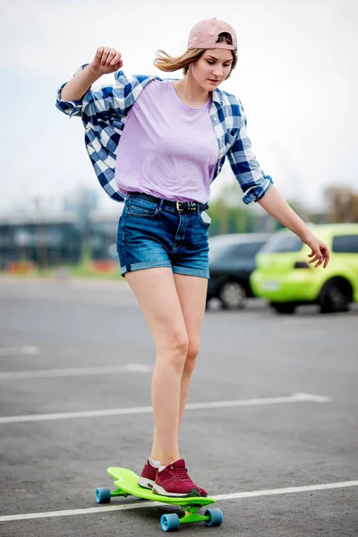 Une belle blonde souriante portant une chemise à carreaux, un bonnet et un short en denim se balance sur le longboard sourire et étire la main. Sport et style cool — Photo