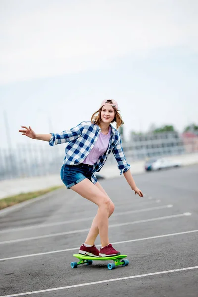 Egy gyönyörű mosolygó szőke lány visel kockás inget, sapka és farmer rövidnadrág longboarding Míg kinyújtva a kezét. Sport-és hidegstílus — Stock Fotó