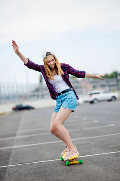 Uma linda garota loira vestindo camisa xadrez, boné e calções jeans é longboard enquanto estende as mãos. Esporte e estilo legal . — Fotografia de Stock