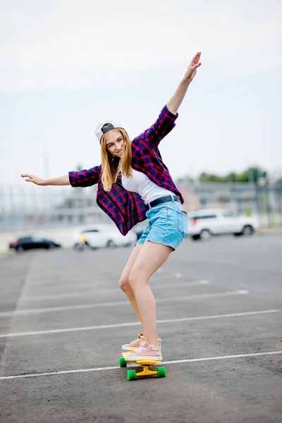 Een mooi blond meisje dat geruite overhemd, pet en denim shorts draagt, is Longboarden terwijl hij haar handen uitreikt. Sport en coole stijl. — Stockfoto