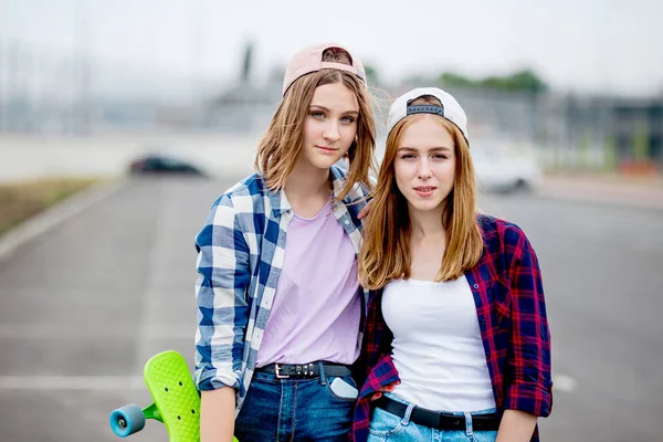 Twee mooie glimlachende blonde meisjes die geruite shirts, caps en denim shorts dragen staan op de lege parkeerplaats met longboards in hun handen. Sport en coole stijl. — Stockfoto