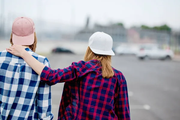 Dwie blond dziewczyny ubrane w kratkę koszule, czapki i szorty jeansowe stoją z plecami na pustym parkingu. Sport i fajny styl. — Zdjęcie stockowe