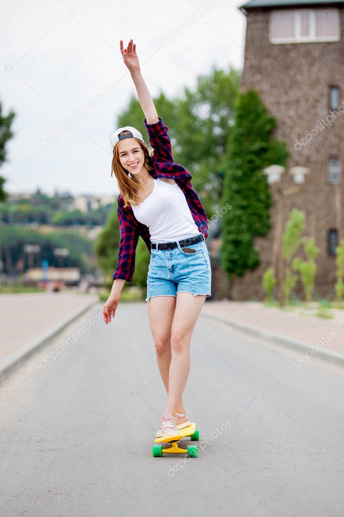 A beautiful smiling blond girl wearing checkered shirt, white cap and denim shorts is balancing on the yellow longboard and stretching up her hand. Sport and cool style