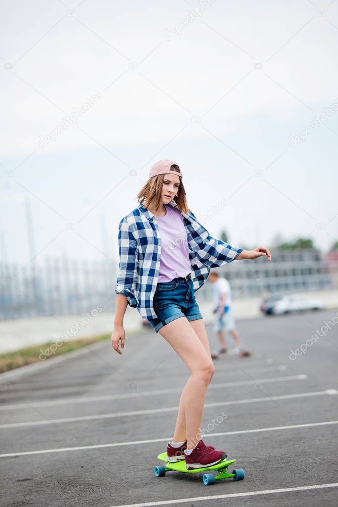 A beautiful blond girl wearing checkered shirt, cap and denim shorts is longboarding while stretching out her hands. Sport and cool style