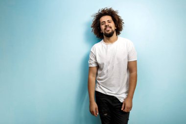 Bir kıvırcık kafalı adam yırtık kot pantolon ve beyaz T-shirt giyen güvenle görünüyor. Mavi üzerinde duran açık fikirli ve kolay olacak adam.