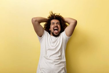 Kıvırcık kafalı brunet adam bağırarak ve kaslı ellerini başının arkasında tutarak. Öfkeli yukarı bak ve sarı zemin üzerine parlak duygular