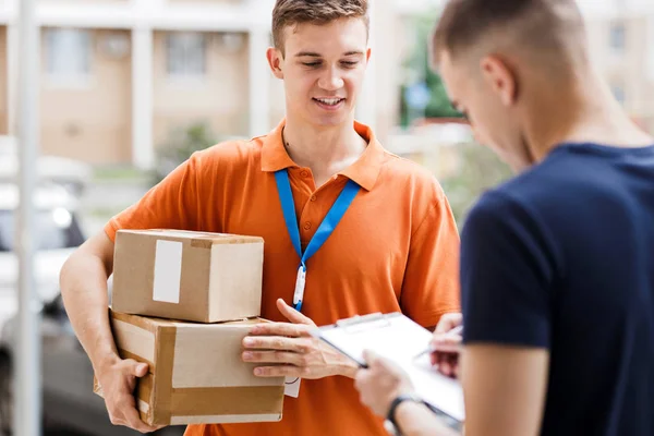 Una persona sonriente que lleva una camiseta naranja y una etiqueta con su nombre está entregando un paquete a un cliente, que está poniendo su firma en el recibo. Trabajador amable, servicio de entrega de alta calidad — Foto de Stock