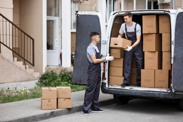 Two young handsome movers wearing uniforms are unloading the van full of boxes. House move, mover service