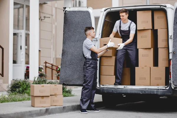 Dos jóvenes mozos guapos con uniformes están descargando la furgoneta llena de cajas. Movimiento de casa, servicio de mudanza — Foto de Stock