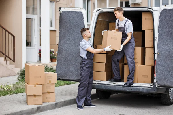 Dwóch młodych przystojny przeprowadzki noszenie mundurów są rozładunku van pełne pudła. Dom przesunąć, mover service — Zdjęcie stockowe