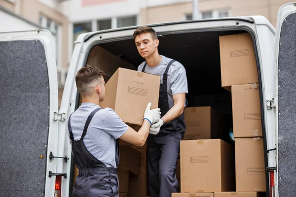 Dos jóvenes mozos guapos con uniformes están descargando la furgoneta llena de cajas. Movimiento de casa, servicio de mudanza — Foto de Stock