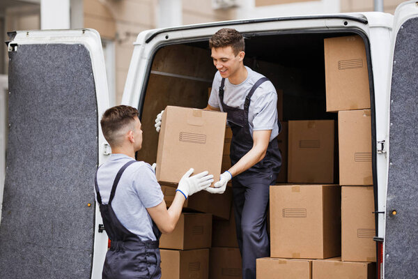 Two young handsome movers wearing uniforms are unloading the van full of boxes. House move, mover service