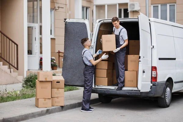 两个穿着制服的年轻帅哥正在卸货的面包车装满箱子。房子移动, 搬运工服务 — 图库照片