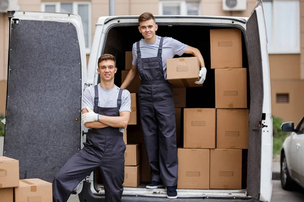 Two young handsome smiling movers wearing uniforms are unloading the van full of boxes. House move, mover service — Stock Photo, Image