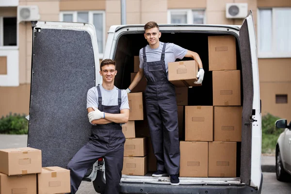 Two young handsome smiling movers wearing uniforms are unloading the van full of boxes. House move, mover service