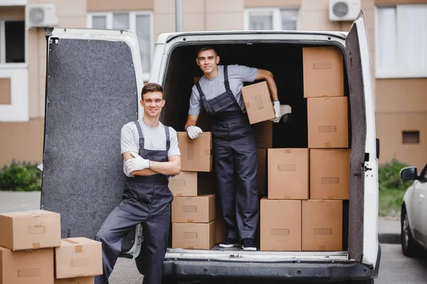 Dos jóvenes mozos guapos y sonrientes que llevan uniformes están descargando la furgoneta llena de cajas. Movimiento de casa, servicio de mudanza — Foto de Stock
