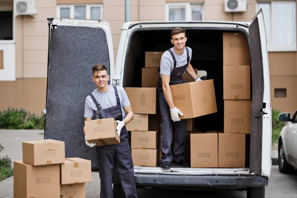 Dos jóvenes mozos guapos y sonrientes que llevan uniformes están descargando la furgoneta llena de cajas. Movimiento de casa, servicio de mudanza — Foto de Stock