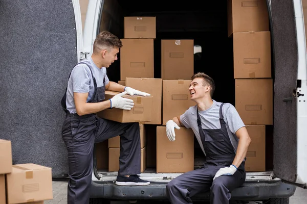 Dos jóvenes mozos guapos que llevan uniformes se ríen mientras descargan la furgoneta llena de cajas. Movimiento de casa, servicio de mudanza — Foto de Stock