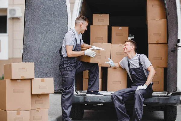 Dos jóvenes mozos guapos que llevan uniformes se ríen mientras descargan la furgoneta llena de cajas. Movimiento de casa, servicio de mudanza — Foto de Stock