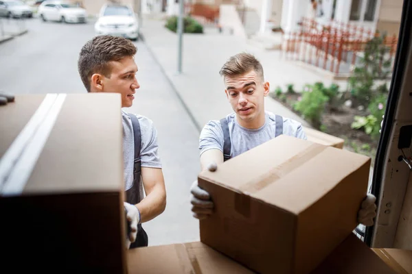 Dwóch młodych przystojny uśmiechający się przeprowadzki noszenie mundurów są rozładunku van pełne pudła. Dom przesunąć, mover service — Zdjęcie stockowe