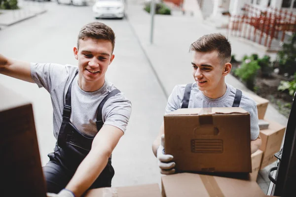 Dois jovens e bonitos homens sorridentes vestindo uniformes estão descarregando a van cheia de caixas. Mudança de casa, serviço de transporte — Fotografia de Stock