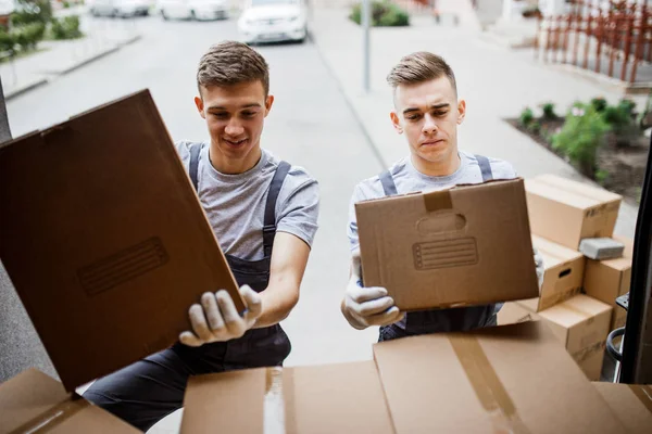 Två unga vackra leende movers bär uniformer lossning van full av lådor. Hus flytta, mover service — Stockfoto