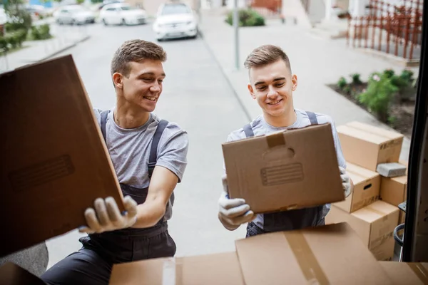 Két fiatal csinos mosolygó mozgatói egyenruhát visel kirakodás a doboz tele van. Ház mozog, mozgató szolgáltatás — Stock Fotó