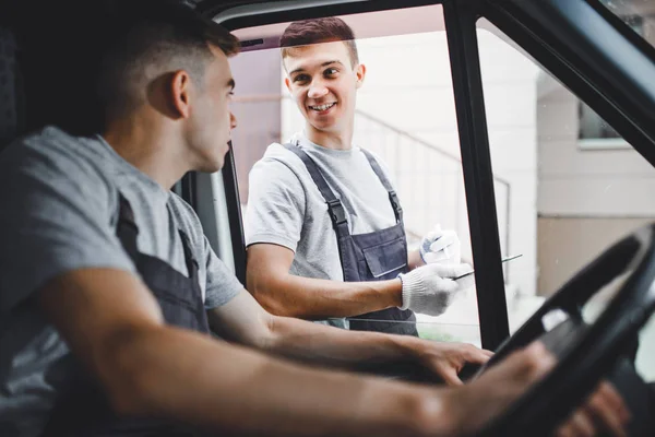 A young handsome guy wearing uniform is looking out of the car window. Another worker wearing uniform is holding the clipboard. House move, mover service — Stock Photo, Image