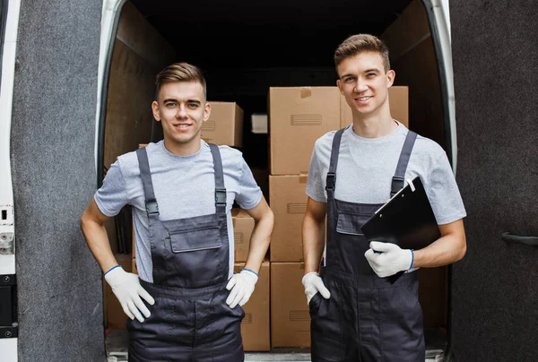 Dos jóvenes y guapos trabajadores sonrientes con uniformes están parados frente a la furgoneta llena de cajas. Movimiento de casa, servicio de mudanza — Foto de Stock