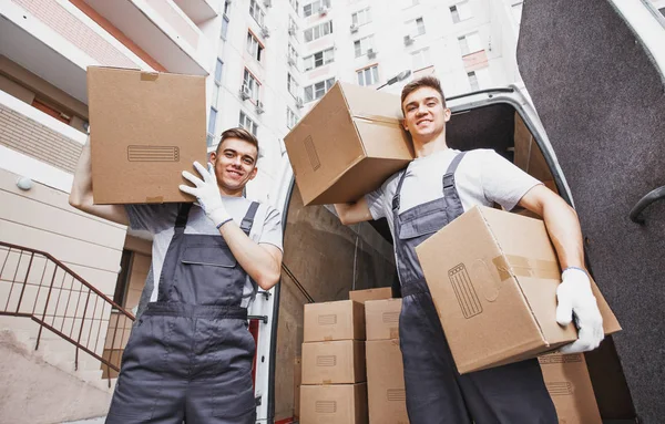 Two young handsome smiling workers wearing uniforms are standing in front of the van full of boxes holding boxes in their hands. House move, mover service — Stock Photo, Image