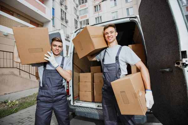 Dos jóvenes y guapos trabajadores sonrientes con uniformes están de pie frente a la camioneta llena de cajas con cajas en las manos. El bloque de pisos está en el fondo. Movimiento de casa, servicio de mudanza — Foto de Stock