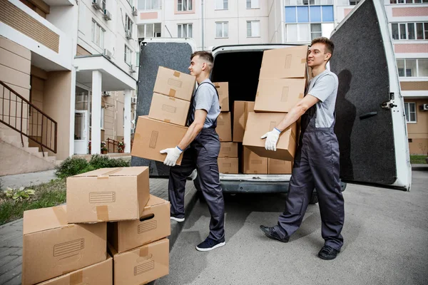 Dois jovens e bonitos trabalhadores sorrindo de uniforme estão descarregando a van cheia de caixas. O bloco de apartamentos está no fundo. Mudança de casa, serviço de transporte — Fotografia de Stock