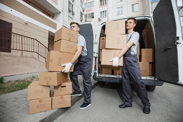 Dois jovens e bonitos trabalhadores sorrindo de uniforme estão descarregando a van cheia de caixas. O bloco de apartamentos está no fundo. Mudança de casa, serviço de transporte — Fotografia de Stock